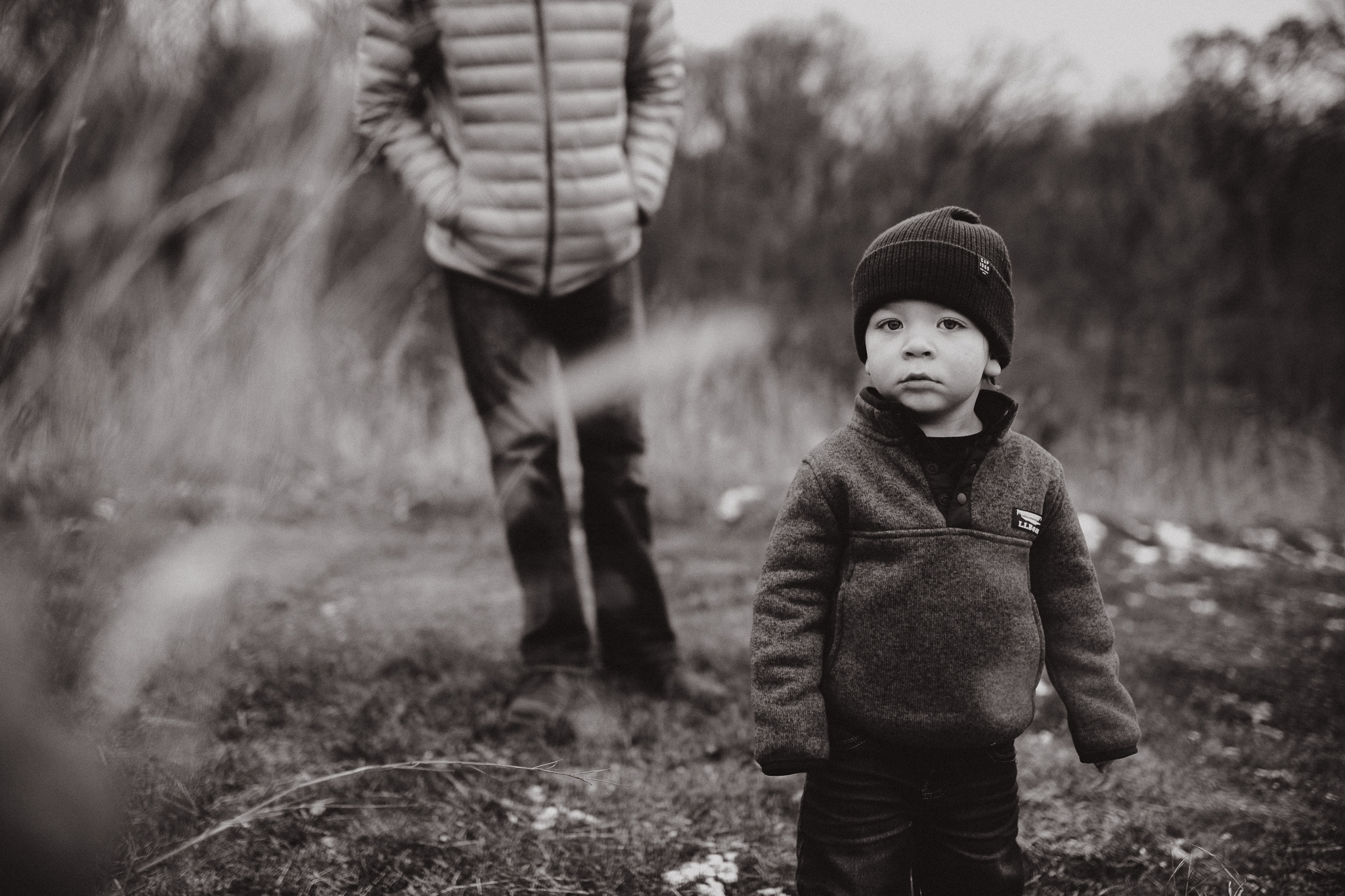 Toddler boy looking through the tall grass as his father stands behind him.